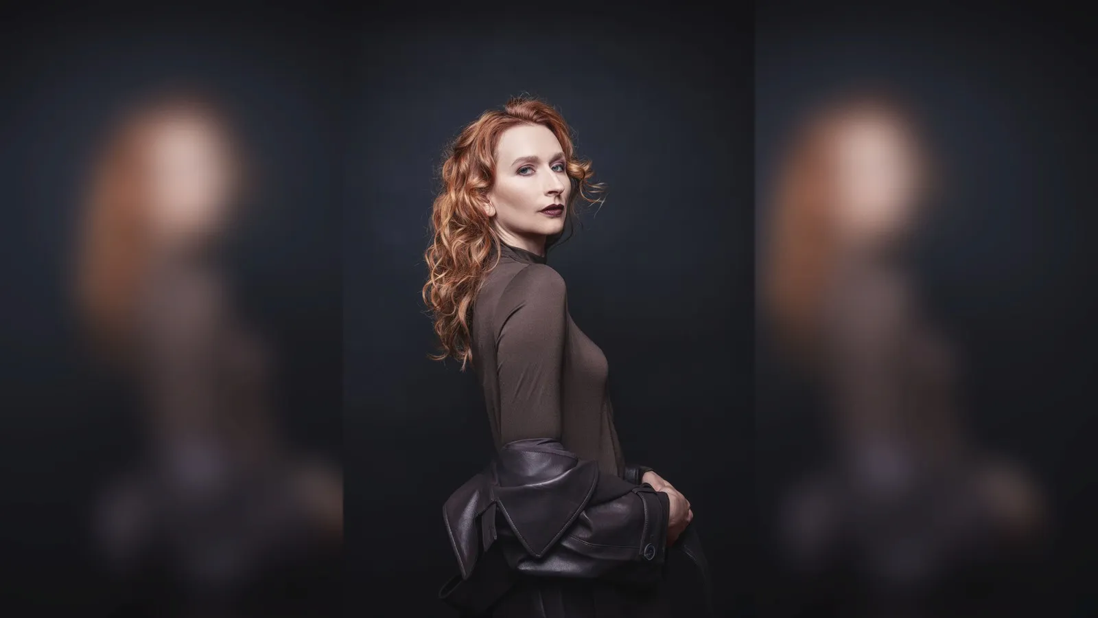 Eine Frau mit kupferroten Locken und dunkelrot-bräunlichem Lippenstift schaut in Richtung Kamera. Sie trägt ein braunes Oberteil sowie eine nur halb angezogene Lederjacke.